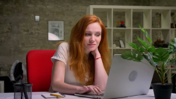 Die kleinlich lächelnde kaukasische Ingwerunternehmerin blickt auf den Laptop-Bildschirm und hält ihren Kopf mit der Hand in einem hellen Büro neben dem Laptop. — Stockvideo