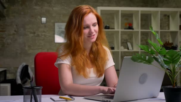 Feliz hermosa sonrisa de chica de jengibre caucásico, sentado en su escritorio y el uso de la computadora portátil durante el trabajo, ser autónomo y satisfecho — Vídeo de stock