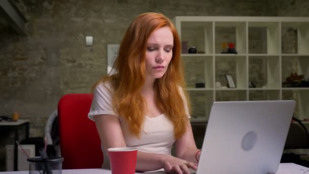 Mujer pelirroja inteligente está escribiendo en su computadora portátil y mirando la pantalla precisamente con confianza, concentrada en su trabajo, interior, fondo de oficina de ladrillo — Vídeos de Stock