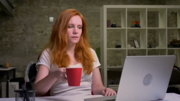 Великолепная рыжая кавказская бизнесвумен только что закончила работу за компьютером, спокойно сидя, расслабившись, попивая из красного кубка un brick office — стоковое видео