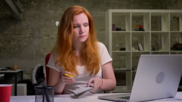 Inteligente mujer de negocios jengibre está mirando a la pantalla del ordenador portátil y escribir algunas notas con cara enfocada seria, sentado en interiores, oficina vibras de trabajo — Vídeo de stock