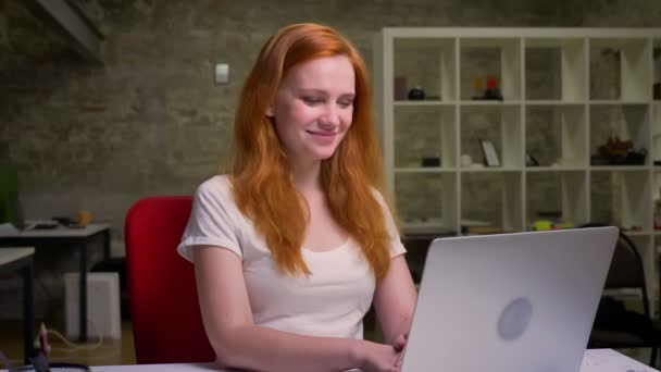 Linda mulher caucasiana artística ruiva de cabelos vermelhos está usando seu laptop enquanto está sentado e sorrindo para a tela feliz, sentado em casa, fundo de tijolo — Vídeo de Stock