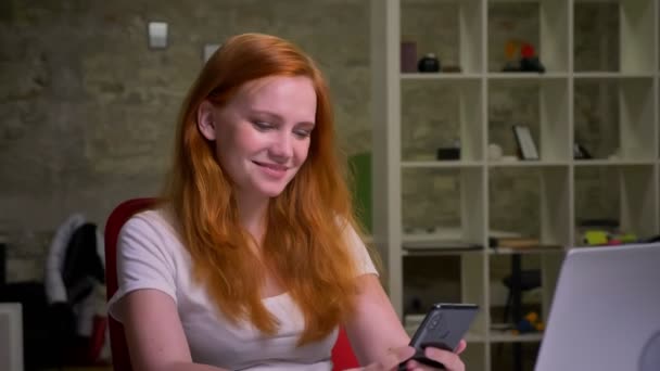 Souriant impressionnant caucasien aux cheveux roux femelle regarde attentivement ordinateur portable et assis dans la chaise, illustration de bureau, intérieur, réalité moderne — Video