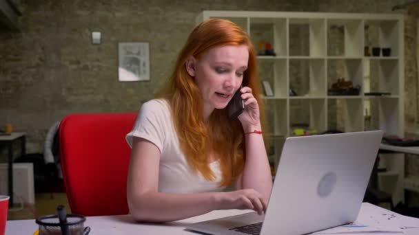 Trabajando chica caucásica está teniendo conversación telefónica mientras está sentada y usando su computadora portátil, ilustración de oficina, mujer enérgica — Vídeo de stock