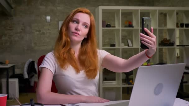 Linda hembra caucásica pelirroja está tomando selfie con la cara concentrada cerca de su computadora portátil en el escritorio, oficina ligera en el fondo — Vídeos de Stock
