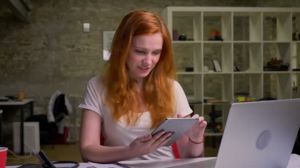 Smiling cute caucasin ginger lady is usinf tablet, looking creative while sitting in light office near computer — Stock Video
