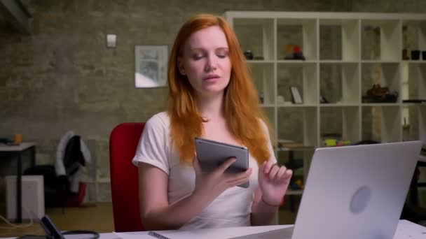 Piacevole felice caucasico dai capelli rossi ragazza è seduta in ufficio isolato e tenendo tablet con schermo verde su di esso, essendo rilassato e freddo, posizione naturale — Video Stock