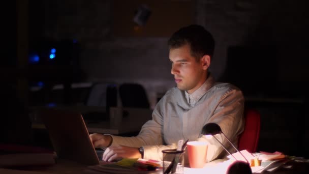 Energetic modern caucasian businessman is typing on keyboard of his laptop while sitting indoor, dark background, late time, empty office — Stock Video