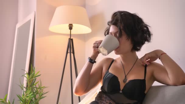 Feliz mujer caucasain sonriente en ropa interior de encaje negro sentado desnudo en el sofá y relajante, beber café y disfrutar de su día, fondo claro interior — Vídeo de stock