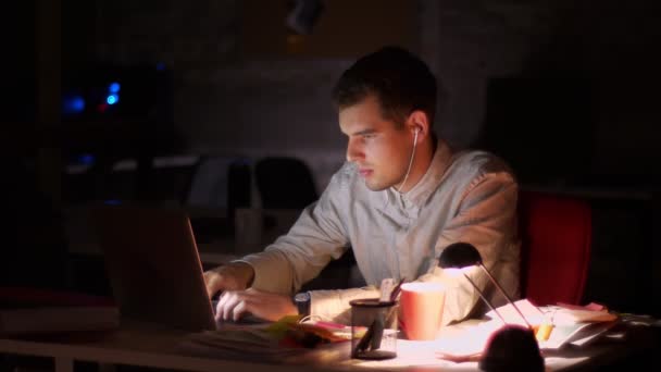 Cute caucasian casual male is listening music in earphones, moving while sitting and typing in laptop productively, relaxed and chill, dark late in office — Stock Video