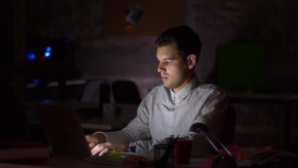 Lindo hombre caucásico está escribiendo en su laprop y bostezar mientras está sentado en la oficina oscura, bclack fondo, productividad e ilustración moderna — Vídeo de stock