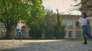 Rahat sevimli dans kızlar üzerinde Square genel yer gösterilen hareketleri ve mutlu bir şekilde gerçekleştirmek, arka plan, güneşli bir gün, güzel sanatlar binalarda
