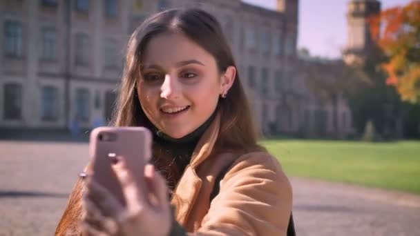 Menina branca sorridente bonito está tomando selfies com sorriso alegre e segurando seu telefone com uma mão enquanto está fora no fundo urbano — Vídeo de Stock