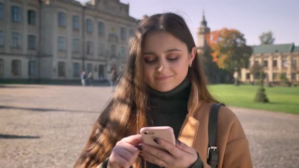 Impresionante hembra caucásica está desplazando su teléfono y haciendo cara divertida después de pensativo serio uno, de pie en el área urbana cerca de edificios antiguos, ilustración soleada . — Vídeos de Stock