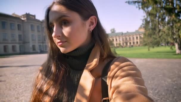 Cute happy caucasian female is holding web camera and close-up and looking at herself while standing outdoor in sunlights, urban background — Stock Video