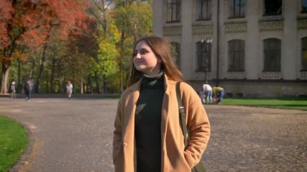 Mooie Kaukasische vrouw loopt in zonnige stedelijk gebied en vawing met schattige glimlach, herfst vibes, groene achtergrond, beweging — Stockvideo