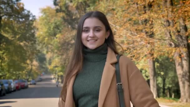 Sorridente femmina caucasica impressionante sta godendo soleggiata giornata autunnale e guardando felicemente la fotocamera, in piedi all'aperto con alberi sullo sfondo, in stile urbano — Video Stock