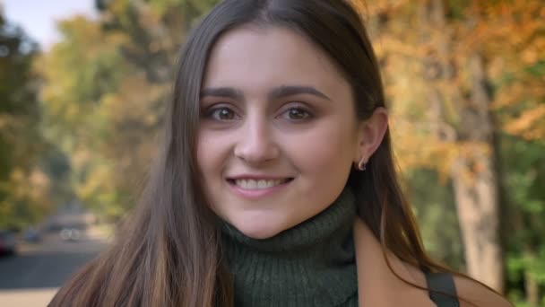 Primer plano hermosa sonrisa en la cara de chica caucásica con increíbles ojos marrones sonriendo y de pie en la calle soleada, cerca del parque, brilla el sol — Vídeo de stock