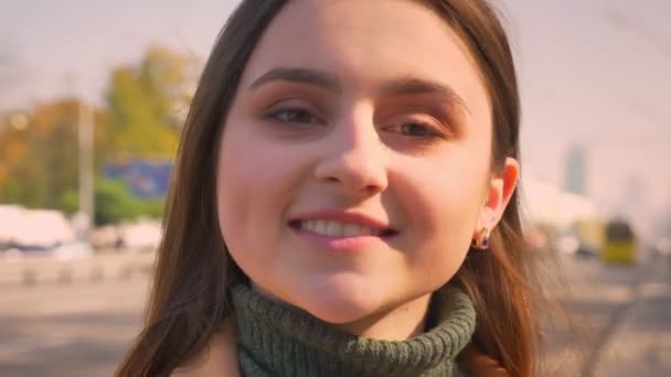 Selbstbewusst lächelnde Kaukasierin blickt präzise in die Kamera, bewegt sich und lächelt kühl, während sie draußen in der Nähe der Autobahn im Sonnenlicht steht — Stockvideo