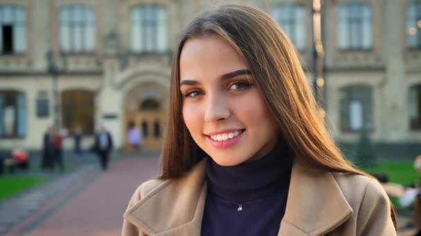 Nahaufnahme Porträt einer lächelnden kaukasischen Frau, die mit glücklichem Gesicht und schönen braunen Augen und langen Haaren direkt in die Kamera blickt, urbaner Hintergrund, sonniger Tag — Stockvideo