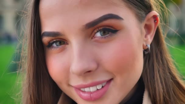 Linda hembra caucásica sonriente con bonitos ojos marrones y hermoso aspecto natural, de pie enfocado y mirando a la cámara relajada, al aire libre, durante el día — Vídeo de stock