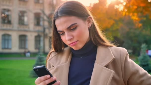 Emocional linda mujer caucásica con hermoso pelo largo está de pie y mirando el teléfono, mientras lo usa con la cara reflexiva, la luz del sol en el fondo, edificios culturales — Vídeos de Stock