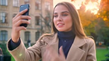Tatlı güzel beyaz kadın telefonunu web kameraya bakıyor ve görüntülü görüşme sahip, üzerinde daire dönüm ve izole üzerinde kentsel arka plan güneşli bir günde hareket