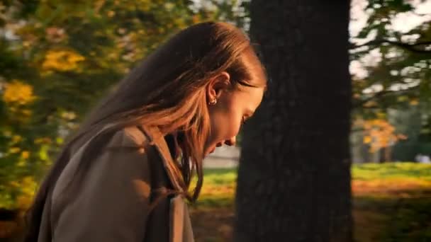 Motion beelden, schitterend Kaukasische meisje in profiel maaien en wandelen in de buurt van de camera in de Gouden herfst park en gelukkig genieten van sunlights op haar gezicht, natuurlijke artistieke afbeelding buiten — Stockvideo