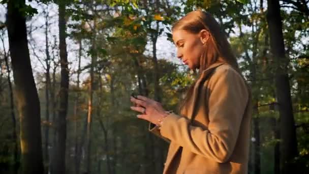 Pretty caucasian female is walking in beautiful fall park and taking pictures of nature, trees as background and happy calm model in motion — Stock Video