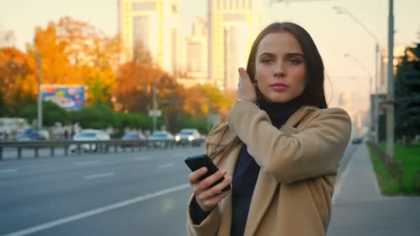 Assez concentré sur le téléphone défilement fille caucasienne est debout près de la route avec des voitures et en attente de transport calmement alors qu'il est fond d'automne et ensoleillé belle journée — Video