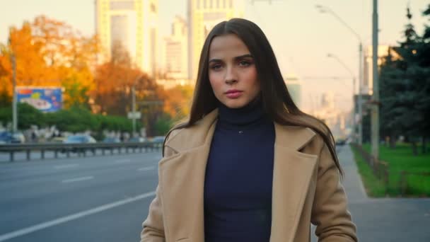 Серьезное лицо красивой кавказской женщины с длинными каштановыми волосами, смотрящей прямо в камеру, стоящей на обочине дороги с движущимся транспортом, осенний день — стоковое видео
