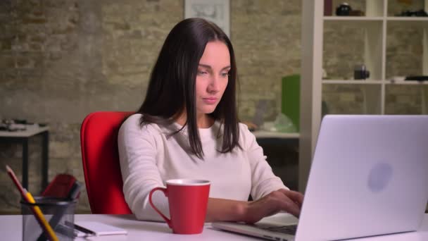 Mulher trabalha no escritório na frente do laptop, bebe café, agita e continua digitando — Vídeo de Stock