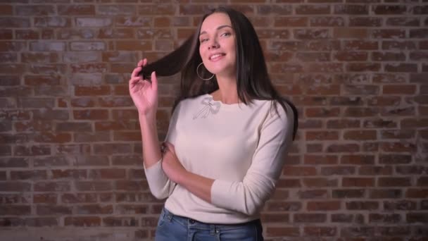 Mujer coqueteando con la cámara y retorciéndose el pelo en el dedo sobre un fondo de ladrillo — Vídeos de Stock