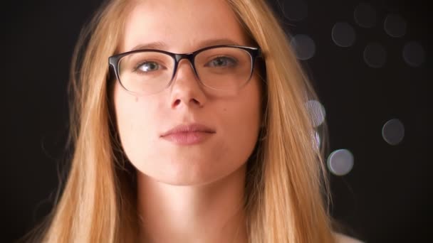 Splendida donna dagli occhiali caucasici bionda è in piedi primo piano e tranquillamente guardando la fotocamera mentre raddrizza gli occhiali con la mano, studio nero al coperto — Video Stock