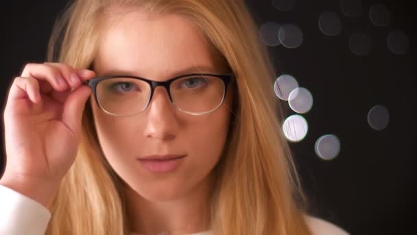 Chica rubia caucásica relajada en gafas con aspecto inteligente, tocando sus gafas con confianza mientras mira a la cámara con cara artística, ilustración de estudio oscuro — Vídeos de Stock