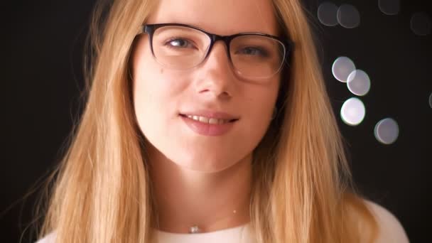 Hübsches Lächeln der kaukasischen Blondine mit Brille blickt kühl in die Kamera, selbstbewusst und entspannt isoliert auf dunklem Hintergrund — Stockvideo
