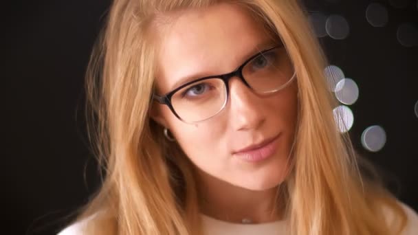 Charmant flirtende kaukasische Blondine steht gerade und blickt selbstbewusst in die Kamera über ihrer Brille, während sie im schwarzen Studio hängt, konzentriert — Stockvideo