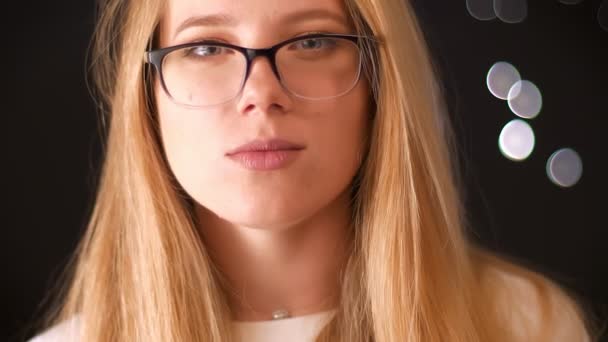 Gorgeous caucasian woman with nice blonde hair puts on reading glasses while standing and looking straight at camera on black background — Stock Video