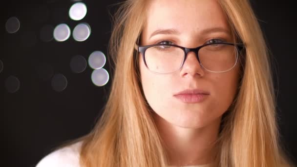 Leuke Kaukasische blonde vrouw in bril is permanent op een zwarte achtergrond en camera kijken met gegarandeerde gezicht, indoor — Stockvideo
