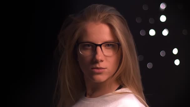 Mignonne femme caucasienne avec de longs cheveux blonds tourne son corps et regarde la caméra précisément à travers des lunettes, tout en se tenant isolé sur fond noir — Video