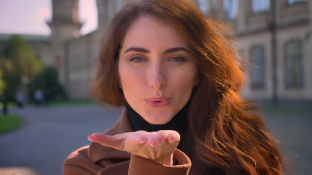 Χαριτωμένο Καυκάσιος μελαχρινή μοντέλο στέκεται έξω στο ηλιοβασιλέματα και closeup στέλνει φυσώντας φιλί στην κάμερα, ψύχρα εικονογράφηση — Αρχείο Βίντεο