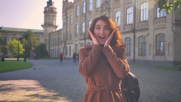 Artistique émotionnelle brune femme caucasienne est la démonstration comme des signes à la caméra et souriant naturellement tout en se tenant debout à l'extérieur sur les bâtiments anciens — Video