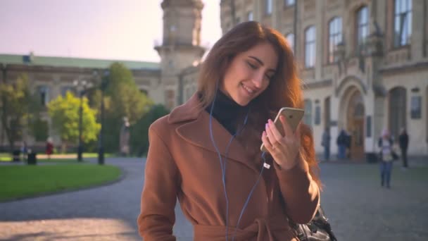 Atrakcyjna brunetka kaukaski jest stały się szczęśliwie i słuchanie muzyki w słuchawki, słoneczny tło miejskie i ilustracja — Wideo stockowe