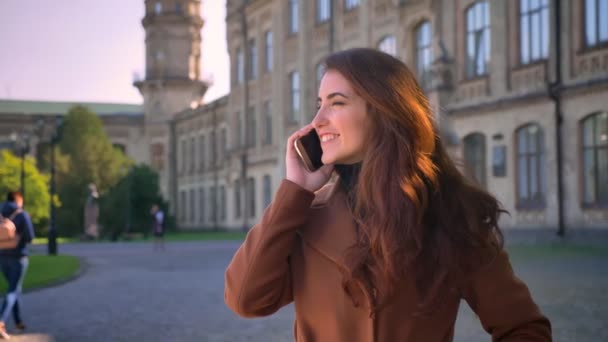 Linda chica morena caucásica está de pie cerca de edificio Antiend y hablando por teléfono con hermosa sonrisa y mirada feliz, al aire libre, las luces del sol — Vídeos de Stock