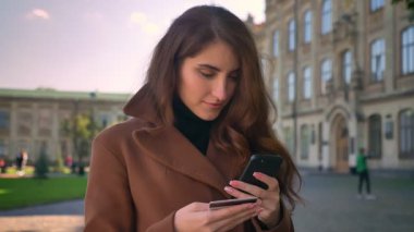 Güzel esmer beyaz kadın ayakta ve onun telefon ve kredi kartı sakince tutarken kameraya bakarak ve kentsel alanda güzel bir güneşli gün harcama