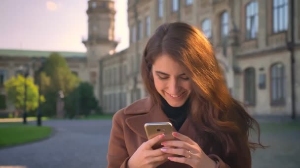 Lächelnde kaukasische Brünette steht und scrollt ihr Handy mit entspanntem Gesicht im Stadtgebiet, sonnige Abbildung, moderne Person — Stockvideo