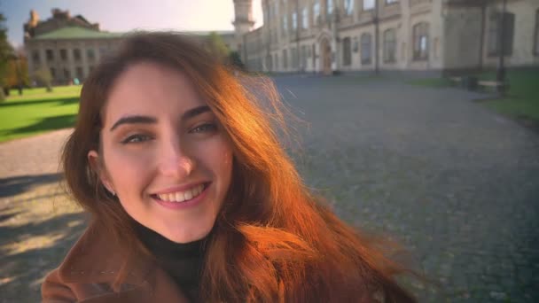 Prachtige brunette staat buiten in zonlicht, houden van de camera en kijken gelukkig met een grote glimlach op haar gezicht, gebouwen op achtergrond — Stockvideo