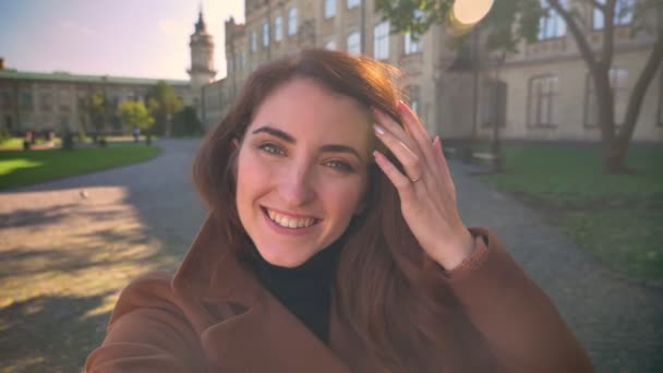 Mujer morena bastante caucásica está sosteniendo la cámara, tomando y saludando durante la conversación web, de pie felizmente bajo los rayos del sol al aire libre, disfrutando de un día soleado — Vídeo de stock