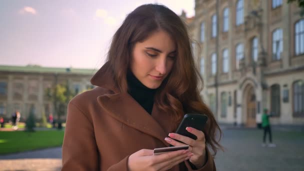 Gyönyörű barna kaukázusi nő állandó és nézi a kamera, miközben a telefon és a hitelkártya nyugodtan és kiadások szép napos városi környezetben — Stock videók