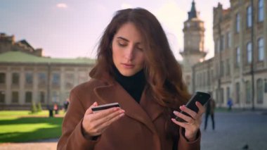 Ayakta beyaz kadın görüntüleri kredi kartı tutan ve vasıl arka planda Bina bilgileri açık havada, güneş ışığı altında dururken kontrol onun telefon numaraları yazmaya.